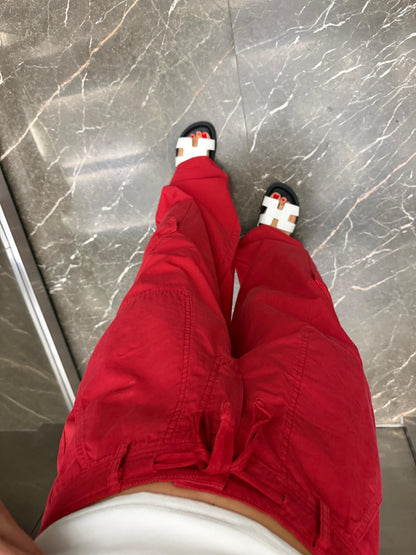 Close-up downward shot of red wide-leg cargo trousers showing thigh cargo pocket detail and wide straight-leg hem, Hermes-style white tan flat sandals with red pedicure, marble elevator floor